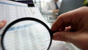 A person is holding a magnifying glass over a piece of paper with numbers on it. The numbers are in different sizes and appear to be related to a financial document